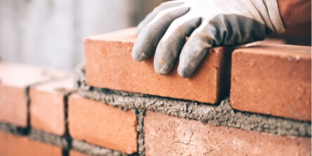 Close-up of a gloved hand placing bricks with mortar in a masonry wall, representing bricklaying and construction work.