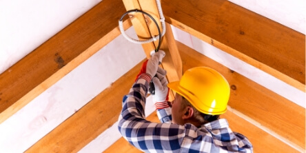 Electrician in a hard hat wiring a ceiling inside a wooden frame building during residential construction.