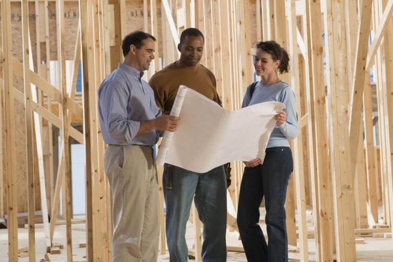 Group of three people, including a couple and a contractor, reviewing blueprints at a construction site with exposed wooden framing, discussing home building plans and design.